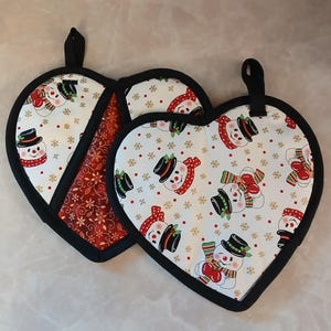 May include: Heart-shaped Christmas potholders with a black trim and hanging loop. The white fabric is patterned with snowmen wearing top hats and scarves. One potholder has a red floral pattern.