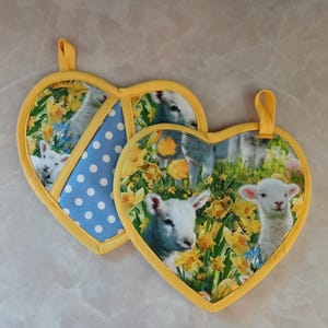 May include: Two heart-shaped pot holders with a yellow border. One pot holder features a blue and white polka dot pattern. The other pot holder features a pattern of lambs in a field of yellow daffodils.