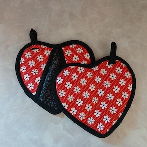 Puede incluir: Dos salvamanteles en forma de corazón. Los salvamanteles son rojos con un estampado floral blanco y ribete negro. Cada uno tiene un lazo negro para colgar. Uno de los salvamanteles tiene un estampado floral en blanco y negro en la parte posterior.