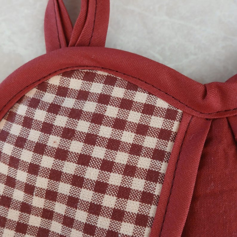 May include: Close-up of a burgundy and cream checkered potholder with a burgundy trim and hanging loop. The potholder has a classic gingham pattern and is made of fabric.