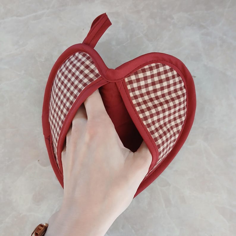 May include: A red and white checkered heart-shaped oven mitt with a loop for hanging. The mitt is made of fabric and has a pocket for the hand. The design is a classic kitchen accessory.