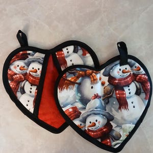May include: Two heart-shaped pot holders with a black trim and a loop for hanging. The pot holders feature a repeating pattern of snowmen wearing red scarves and hats. One pot holder has a red interior.