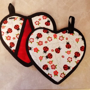 Puede incluir: Dos agarraderas de olla en forma de corazón con ribete negro. Una es roja y la otra es blanca con un patrón de mariquitas rojas y flores blancas.