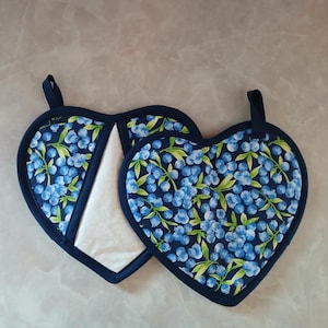 Könnte beinhalten: Zwei herzförmige Topflappen mit einem Blaubeer-Druck. Die Topflappen sind marineblau mit weißem Hintergrund und haben ein weißes gestepptes Futter.