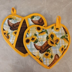 May include: Two heart-shaped pot holders with a yellow border and a white background. The pot holders feature a pattern of sunflowers and cowboy boots.