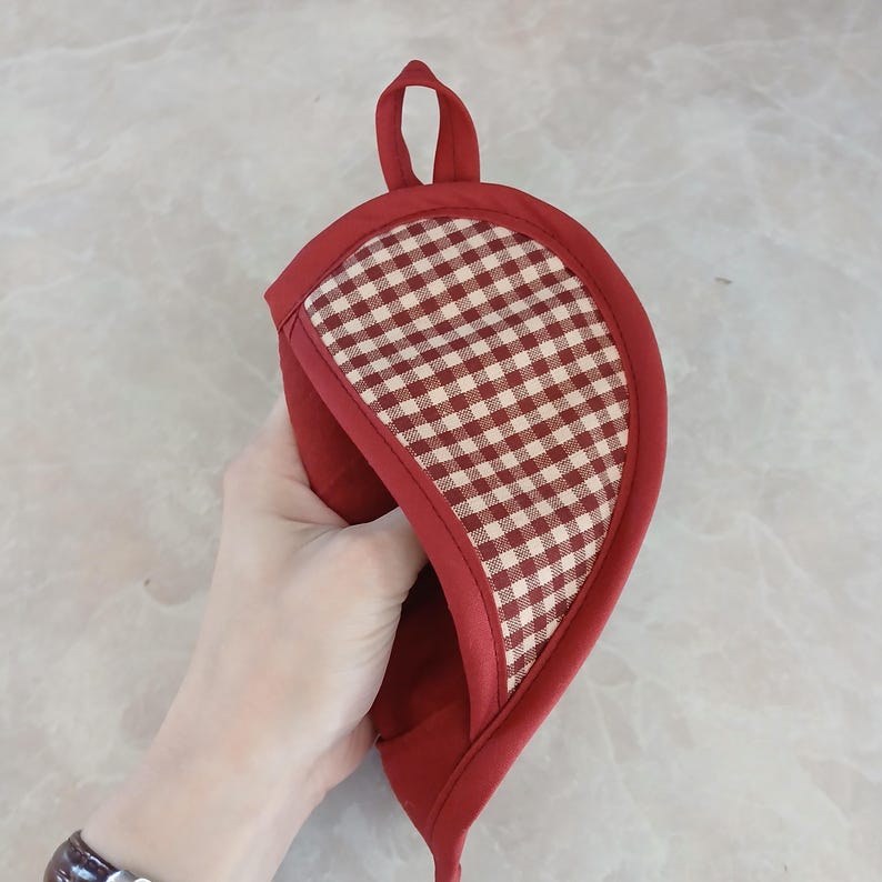 May include: Red and white checkered oven mitt with a loop for hanging. The mitt is red with a checkered pattern on the front and a red trim. The mitt is held up by a hand.