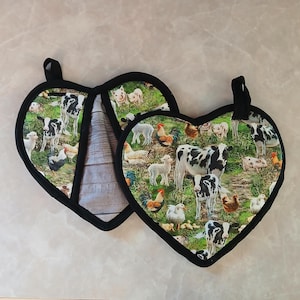 Puede incluir: Dos agarraderas de olla en forma de corazón con ribete negro. Las agarraderas tienen un fondo verde con un estampado de animales de granja de vacas, cerdos, pollos y corderos.