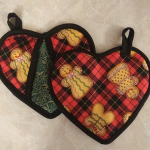 以下が含まれることがあります： 黒い縁と吊り下げループが付いた、ハート型の鍋つかみ2つ。鍋つかみは、ジンジャーブレッドマンのデザインが施された赤と黒のチェック柄です。1つの鍋つかみは、内側に緑色の模様の生地が付いています。