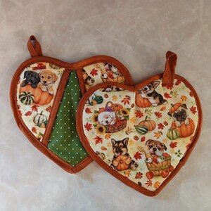 May include: Two heart-shaped pot holders with a brown trim. The pot holders feature a green and white polka dot fabric and a cream fabric with a pattern of puppies, pumpkins, and fall leaves.