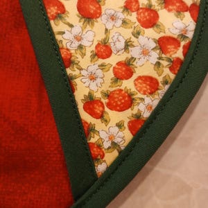 Pu&ograve; includere: Primo piano di un bordo di tessuto verde con uno sfondo di tessuto giallo che presenta un motivo ripetuto di fragole rosse e fiori bianchi.