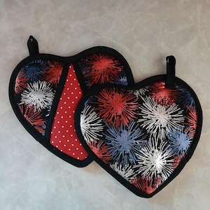 May include: Two heart-shaped pot holders with black trim. One features a red and white polka dot pattern, while the other has a black background with red, white, and blue firework-like designs. Each has a black loop for hanging.