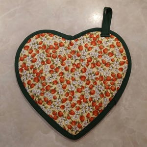 Pu&ograve; includere: Un sottopentola a forma di cuore con un bordo verde e uno sfondo giallo con un motivo di fragole rosse e fiori bianchi.