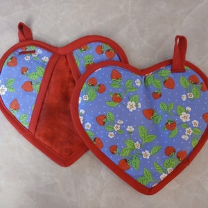 May include: Two red and blue heart-shaped pot holders with a strawberry and flower pattern. The pot holders have a red trim and a loop for hanging.