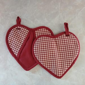 May include: Two heart-shaped oven mitts. The mitts are red with a white and red gingham pattern on the front and a solid red back. Each mitt has a red loop for hanging.