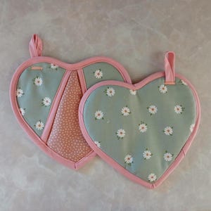 May include: A pair of heart-shaped oven mitts. One mitt is pink with white polka dots and a green floral pattern. The other mitt is green with a white floral pattern. Both mitts have pink trim.