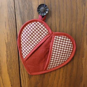 May include: Heart-shaped red and white gingham patterned pot holder with a solid red center. The pot holder is hanging from a dark metal knob on a wooden surface. The pot holder is a kitchen accessory.