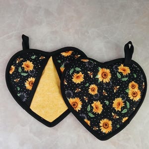 May include: Two heart-shaped pot holders with a black background, yellow sunflowers, and white polka dots. The pot holders have a black border and a loop for hanging. One pot holder is open, revealing a yellow patterned interior.