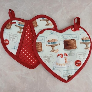 May include: Two red heart-shaped oven mitts with a white background and a blue and brown pattern of baked goods. The mitts have red polka dots on the inside and the text "Premium Baked Goods" and "Fresh Baked" on the outside.
