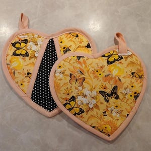 May include: Two heart-shaped pot holders with a floral pattern and butterfly accents. The pot holders are yellow with black butterflies and have a pink trim.
