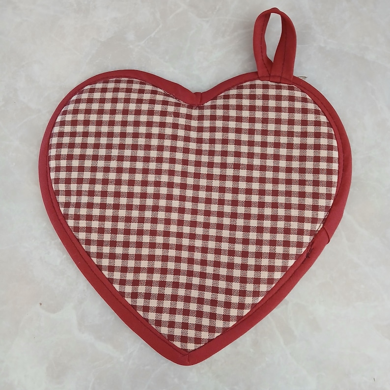 May include: A heart-shaped red and white checkered pot holder with a red border and hanging loop. The pot holder is made of fabric and is designed to protect surfaces from hot cookware.