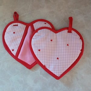 May include: Two heart-shaped pot holders with red trim and a gingham pattern in white and light grey. Small red hearts are scattered across the gingham fabric. Each pot holder has a red loop for hanging.