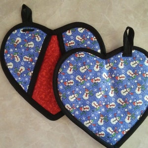 May include: Two heart-shaped pot holders with a snowman pattern on a blue background. One pot holder has a red background with a black border. Both pot holders have black hanging loops.