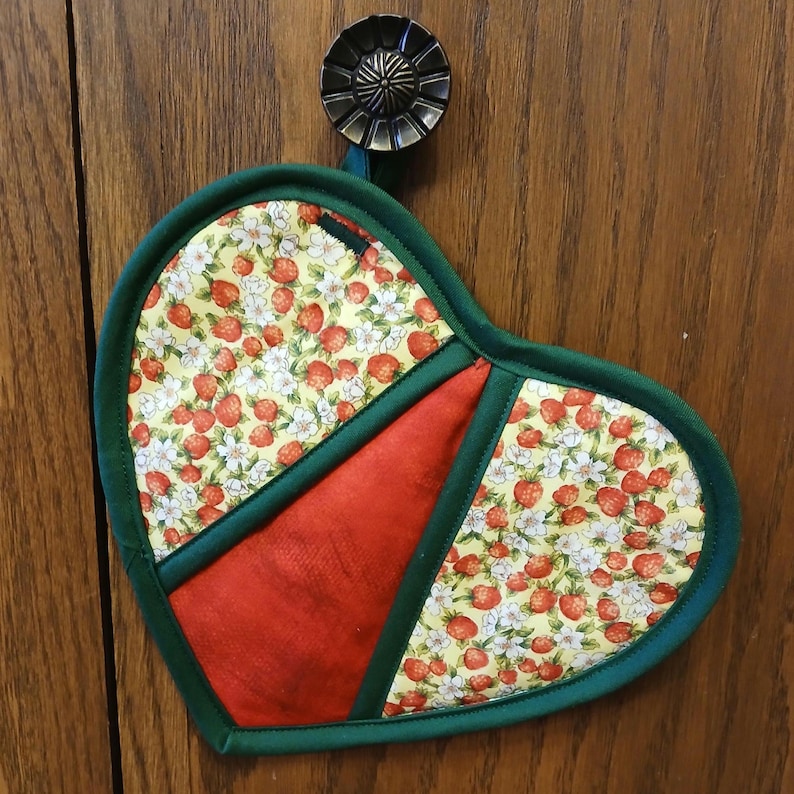 Pu&ograve; includere: Un guanto da forno a forma di cuore verde con un design in tessuto rosso e giallo con fragole e fiori bianchi. Il guanto da forno &egrave; appeso a un gancio.