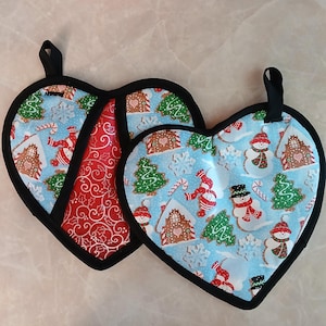 May include: Two heart-shaped pot holders with a winter holiday theme. One pot holder is red with a swirling pattern and the other is blue with a snowman and gingerbread house pattern.