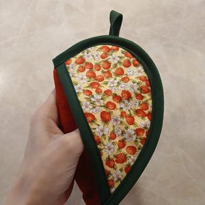 Pu&ograve; includere: Un guanto da forno verde e rosso con un motivo a fragole e fiori. Il guanto da forno ha la forma di una foglia e ha un anello per appenderlo.