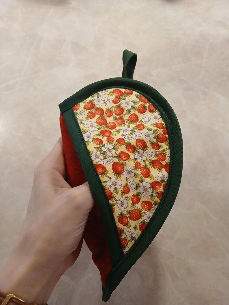 Pu&ograve; includere: Un guanto da forno verde e rosso con un motivo a fragole e fiori. Il guanto da forno ha la forma di una foglia e ha un anello per appenderlo.