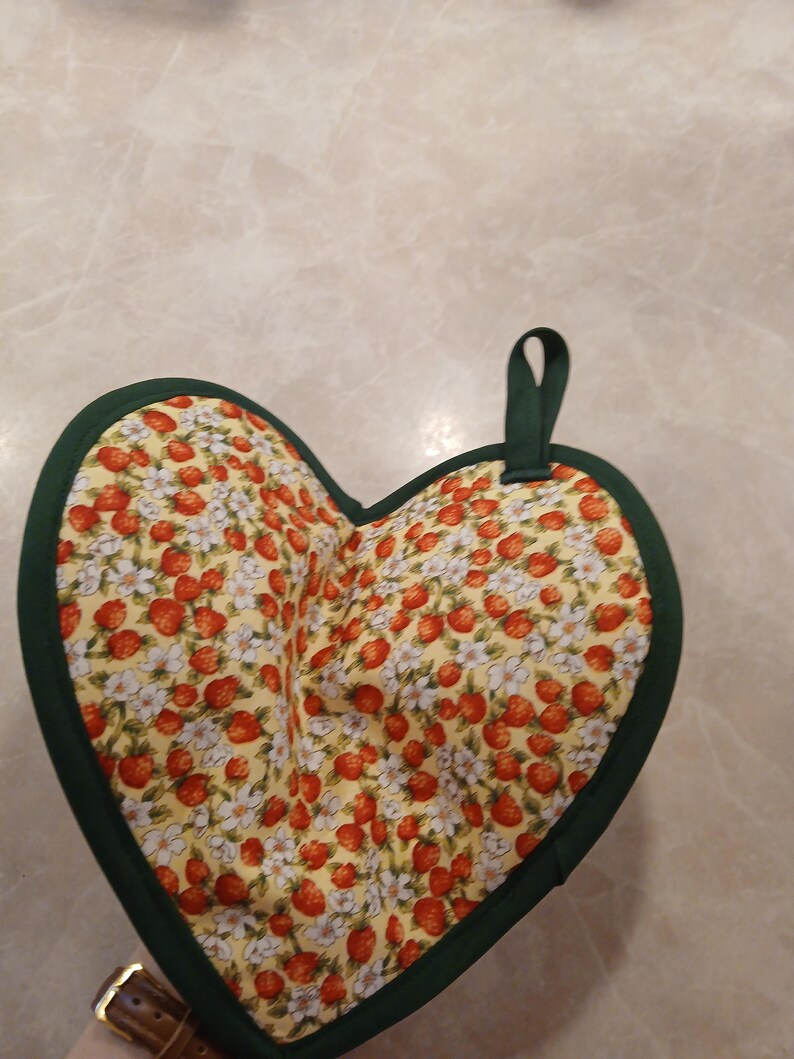 Pu&ograve; includere: Un sottopentola a forma di cuore con un bordo verde e uno sfondo giallo con un motivo di fragole e fiori.