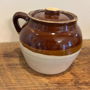 May include: A brown and white ceramic crock with a lid. The crock has a handle and a band of tan color separating the white base from the brown upper portion. The lid has a wooden knob.