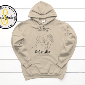 May include: A beige hoodie with a drawstring hood and a front pocket. The design includes a black line drawing of a person with a plant and the text "PLANTS ARE MY SOIL MATES". A circular logo with text is in the upper left corner.