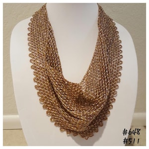 May include: A handmade, open-weave necklace in shades of gold and bronze, displayed on a white mannequin. The intricate design features a scalloped edge and a draped, scarf-like appearance. The image includes the numbers #648 and #511.