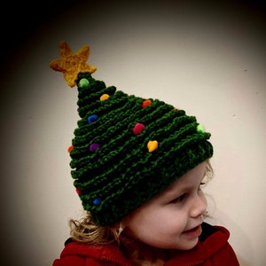 May include: A child wearing a green knitted Christmas tree hat with a yellow star on top and colorful pom-poms. The hat has a green and white striped lining.