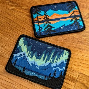 May include: Two rectangular embroidered patches with black borders. One patch depicts a sunset landscape with orange and blue mountains and pine trees. The other patch shows a night scene with green aurora borealis over blue mountains.