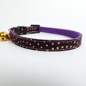 Puede incluir: Collar de gato morado con un patr&oacute;n de estrellas doradas y una campana dorada.