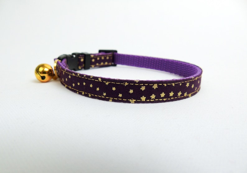Puede incluir: Collar de gato morado con un patr&oacute;n de estrellas doradas y una campana dorada.