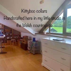 Puede incluir: Una c&oacute;moda blanca con tres cajones y tiradores plateados. La c&oacute;moda est&aacute; en una habitaci&oacute;n con una ventana que da a un campo verde. El texto "Kittyboo collars Handcrafted here in my little studio in the Welsh countryside" es visible en la imagen.