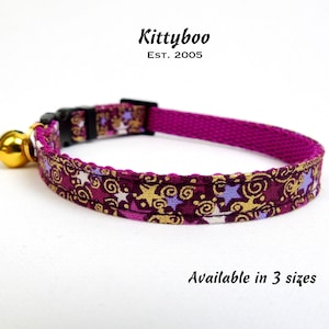 May include: A pink cat collar with a gold and purple star pattern. The collar has a bell attached and the text "Kittyboo Est. 2005" is printed above the collar. The text "Available in 3 sizes" is printed below the collar.