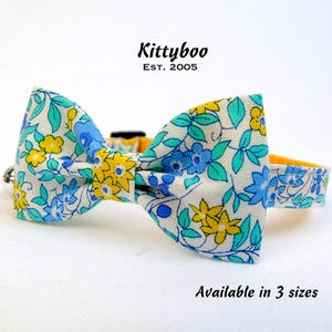 May include: A white, blue and yellow floral patterned bow tie for a pet. The bow tie is attached to a yellow collar. The text "Kittyboo Est. 2005" is above the bow tie. The text "Available in 3 sizes" is below the bow tie.