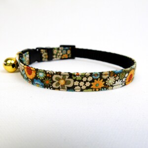 May include: A black breakaway cat collar with a floral pattern in shades of green, blue, orange, and white. The collar has a gold bell attached.