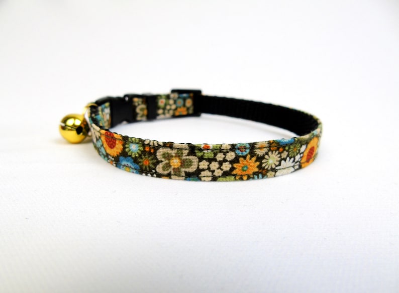 May include: A black breakaway cat collar with a floral pattern in shades of green, blue, orange, and white. The collar has a gold bell attached.
