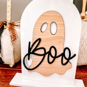 May include: A white wooden ghost with black eyes and the word "Boo" in black lettering. The ghost is on a white stand.