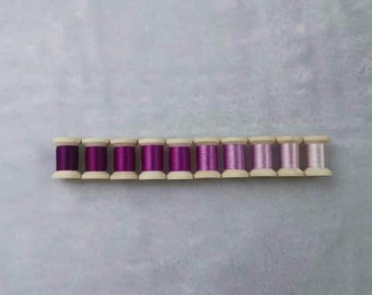 May include: A row of 10 spools of thread in various shades of purple and pink, arranged in a gradient from dark purple to light pink.