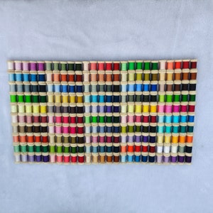 May include: A collection of 100 spools of thread in various colors, arranged in rows on a white surface. The spools are made of wood and have a small hole in the center for threading.