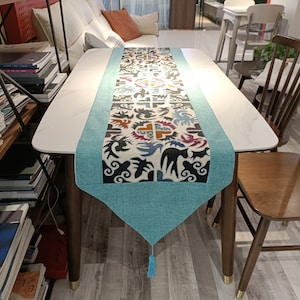 May include: A turquoise table runner with a colourful geometric pattern and tassels. The runner is on a white table with wooden legs and two wooden chairs.