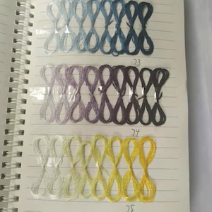 Könnte beinhalten: Ein Spiralnotizbuch, das auf einer Seite mit Stickgarnmustern geöffnet ist. Das Garn ist in einer Reihe von Acht-Muster angeordnet und zeigt eine Reihe von Farben von Hellblau bis Dunkelblau, Lavendel bis Dunkelviolett und Hellgelb bis Gold. Die Nummern 73, 74 und 75 sind sichtbar.