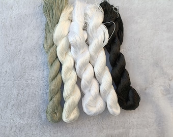 May include: Four twisted hanks of embroidery thread arranged vertically. The colors include a muted sage green, a pale cream, a bright white, and a deep black. Each hank features a smooth, lustrous texture with visible individual fiber strands.
