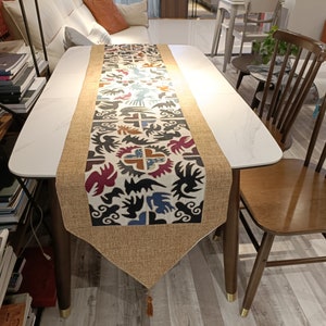 May include: A white table with a brown and beige table runner featuring a colourful geometric pattern. The table runner has a tassel at the end. Two wooden chairs with brown seats are next to the table.